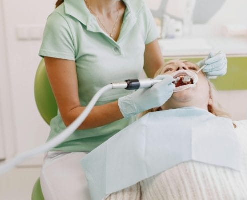 Dental professional in mint scrubs and gloves works inside a patient’s open mouth with dental instruments.