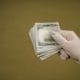 Gloved hand holding a fan of US $100 bills, showing cash in a bundle.
