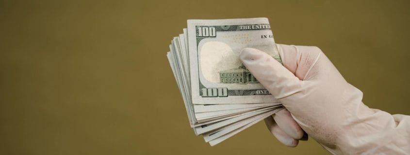 Dental prevention saves money - Unique Dental of Framingham Gloved hand holding a fan of US $100 bills, showing cash in a bundle.