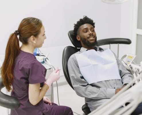 Black man reclining in a dental chair, talking with a female dentist in purple scrubs in a clean clinic.