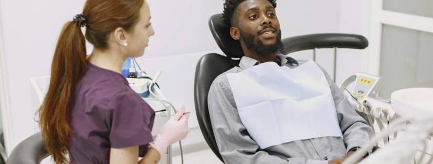 Dentist generations family care - Unique Dental of Framingham Black man reclining in a dental chair, talking with a female dentist in purple scrubs in a clean clinic.