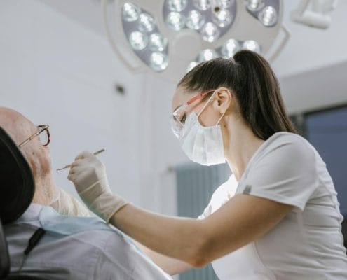Dental professional wearing gloves, a mask, and protective eyewear leans over a patient in a dental chair, performing a procedure under bright overhead surgical lights in a clinic.