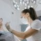 Dental professional wearing gloves, a mask, and protective eyewear leans over a patient in a dental chair, performing a procedure under bright overhead surgical lights in a clinic.