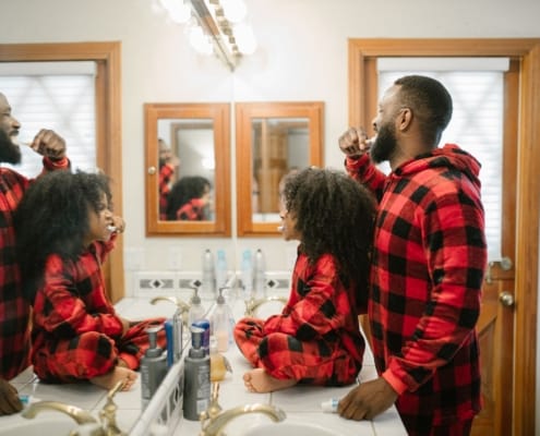 Two men with beards in red plaid shirts brushing their teeth at a bathroom sink, facing mirrors.
