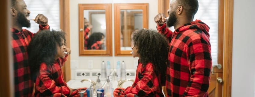 Family brushing teeth healthy habits - Unique Dental of Framingham Two men with beards in red plaid shirts brushing their teeth at a bathroom sink, facing mirrors.