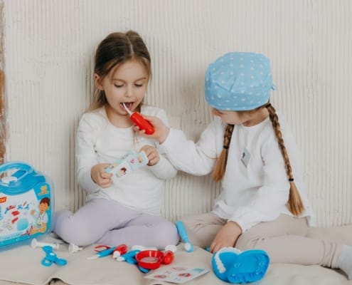 Two young girls play dentist, one brushes the other's teeth with a red toy toothbrush beside a dental kit on the floor.