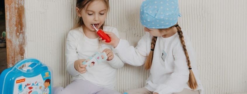 Family dentist children smiling - Unique Dental of Framingham Two young girls play dentist, one brushes the other's teeth with a red toy toothbrush beside a dental kit on the floor.