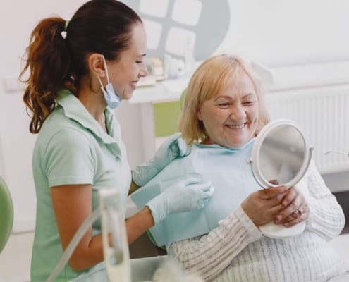 Dental assistant with mask lowered chats and smiles at an elderly patient seated in a dental chair, holding a handheld mirror.