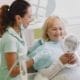 Complete Guide Framingham Family Dentist - Unique Dental of Framingham Dental assistant with mask lowered chats and smiles at an elderly patient seated in a dental chair, holding a handheld mirror.