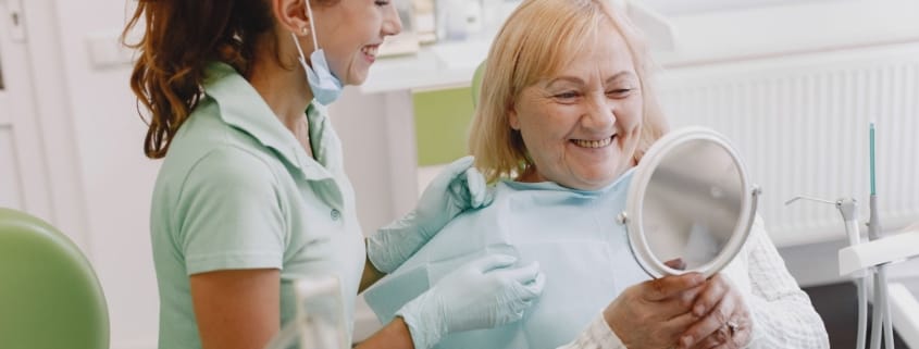 Complete Guide Framingham Family Dentist - Unique Dental of Framingham Dental assistant with mask lowered chats and smiles at an elderly patient seated in a dental chair, holding a handheld mirror.