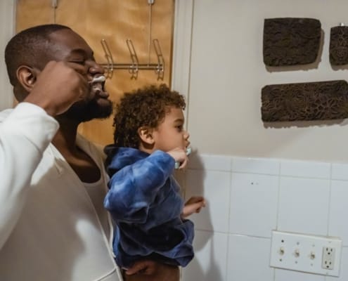 Man holds a young child up to brush the child’s teeth in a bathroom.