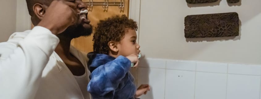 Framingham Family Dentist Makes Dental Care Easy - Unique Dental of Framingham Man holds a young child up to brush the child’s teeth in a bathroom.
