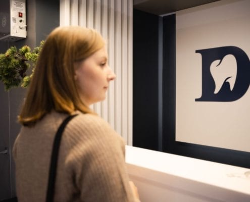 Profile of a woman in a beige sweater standing at a white reception desk with a large blue logo on the wall behind her