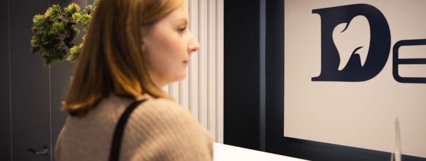 Trusted Framingham Family Dentist - Unique Dental of Framingham Profile of a woman in a beige sweater standing at a white reception desk with a large blue logo on the wall behind her