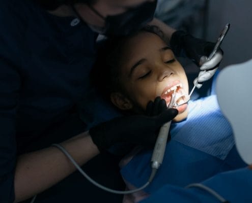 Child patient receiving dental care while a dentist examines her mouth with tools and a mirror, wearing a blue bib and gloves