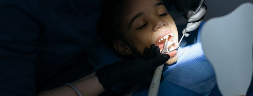 Preventive Care Framingham Family Dentist - Unique Dental of Framingham Child patient receiving dental care while a dentist examines her mouth with tools and a mirror, wearing a blue bib and gloves