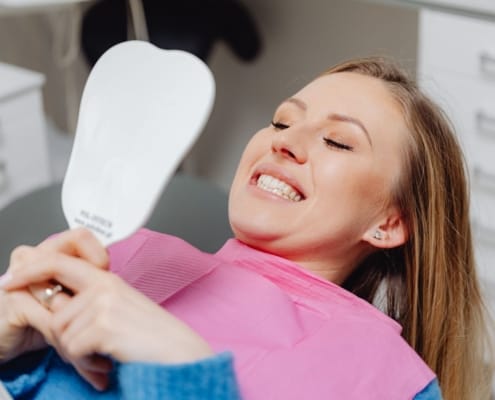 Woman reclined in a dental chair wearing a pink bib, smiling while looking at a handheld mirror