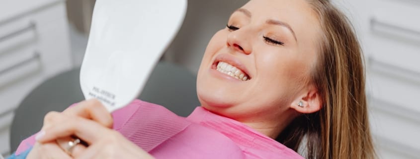 Happy dental visit positive experience - Unique Dental of Framingham Woman reclined in a dental chair wearing a pink bib, smiling while looking at a handheld mirror