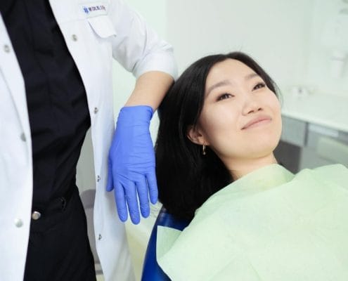 Female patient lying in a dental chair while a clinician in a white coat and blue gloves handles her head.