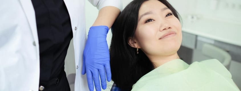 Oral health wellness dental checkup - Unique Dental of Framingham Female patient lying in a dental chair while a clinician in a white coat and blue gloves handles her head.